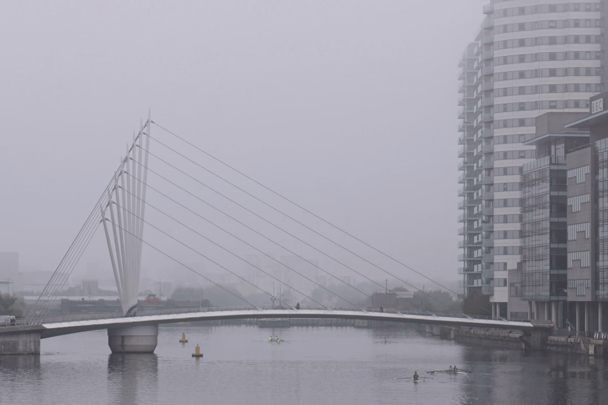 Pasarela atirantada sobre un río en un día de niebla, con edificios difusos al fondo